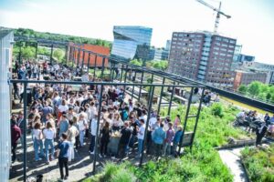 Drukke borrel op een zonnig dakterras aan de Johan Huizingalaan met uitzicht op kantoorgebouwen.